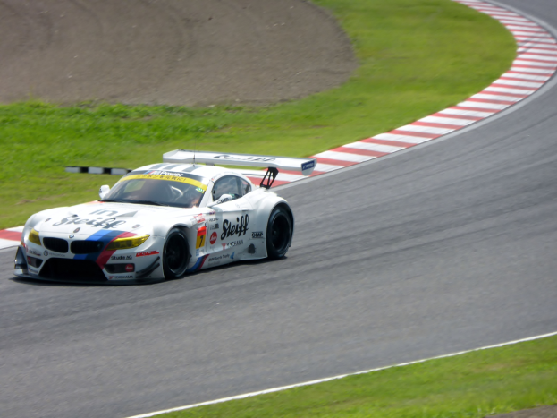 Ein BMW Z4 GT3 Rennauto ist auf der Nürburgring 24 Stunden-Rennstrecke positioniert, umgeben von grünem Gras, mit der Strecke, die sich vor ihm erstreckt.
