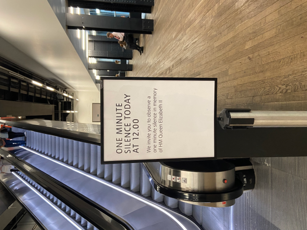 Eine Rolltreppe in einem Flughafen mit einem Schild, auf dem "Eine Minute Stille heute" steht, ein paar Menschen darauf und Lichter an der Decke im Hintergrund.