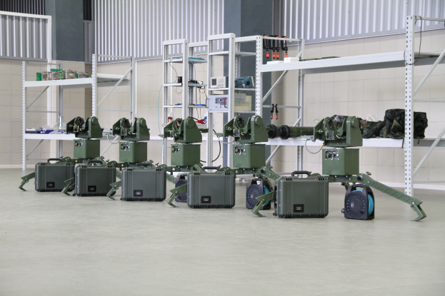 Eine Gruppe von Milit├Ąrrobotern, die auf einem Boden mit Koffern und anderen Gegenst├Ąnden auf umliegenden Gestellen positioniert sind, vor einer Wand mit einem Gel├Ąnder im Hintergrund.