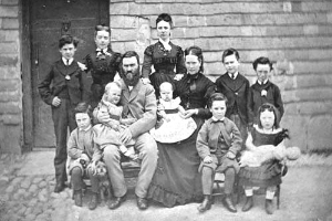 Schwarzes und weißes Foto einer Familie, die vor einem Haus posiert, einige sitzen auf einer Bank und andere stehen, mit einer Wand und einer Tür im Hintergrund.