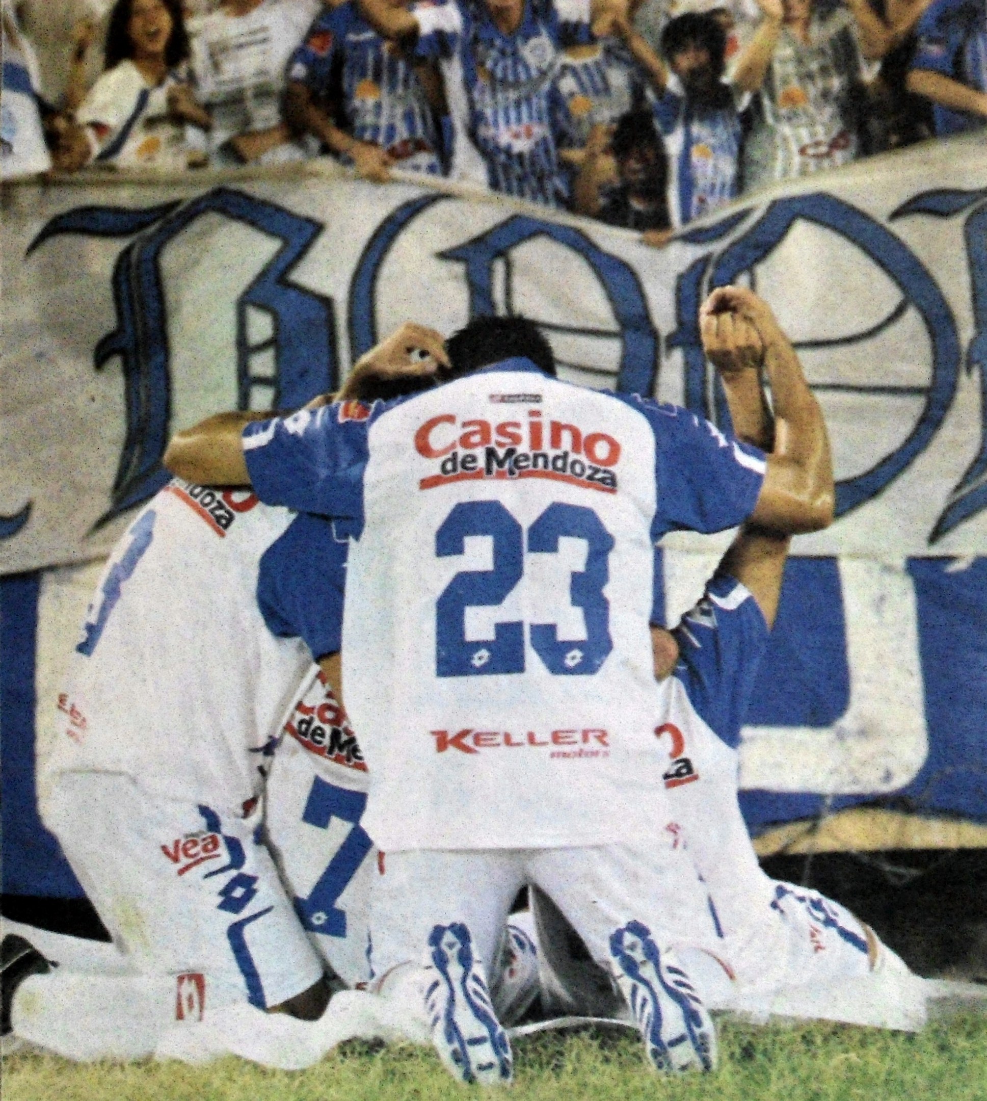 Gruppe von Fußballspielern feiert auf dem Boden vor einer jubelnden Menge, mit einem Banner mit Text im Hintergrund.