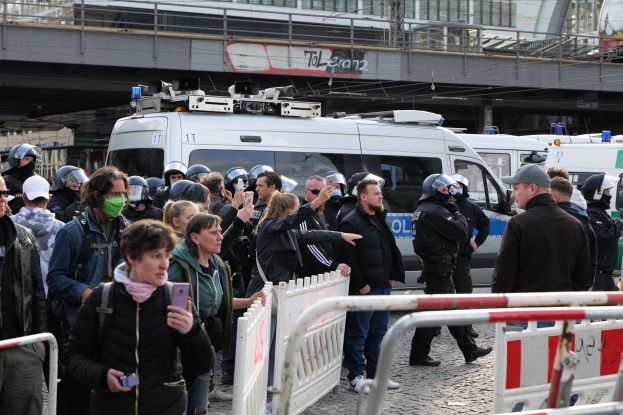 Eine Gruppe von Menschen steht vor Polizeiwagen, einige tragen Helme und halten Telefone, mit Barrikaden im Vordergrund und einer Brücke und Gebäuden im Hintergrund während einer Protestauflösung in Berlin, Deutschland.
