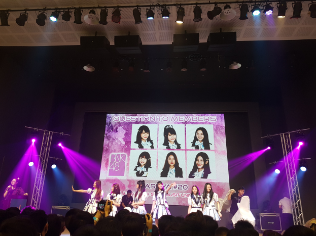 Eine Gruppe von Menschen performs auf einer hell beleuchteten Bühne mit Trägern, mit einem Sänger mit Mikrofon auf der linken Seite und einem Musiker, der ein Instrument spielt, während eine Menge mit Kameras unten zuschaut und ein großer Bildschirm Bilder der K-Pop-Gruppe BK48 anzeigt.