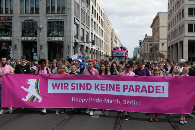 Eine Gruppe von Menschen geht eine Straße in Berlin, Deutschland, entlang und hält eine pinkfarbene Fahne mit der Aufschrift "Happy Pride March" hoch, während Gebäude, Laternenpfähle und Verkehrszeichen die Straße säumen und der Himmel bewölkt ist.