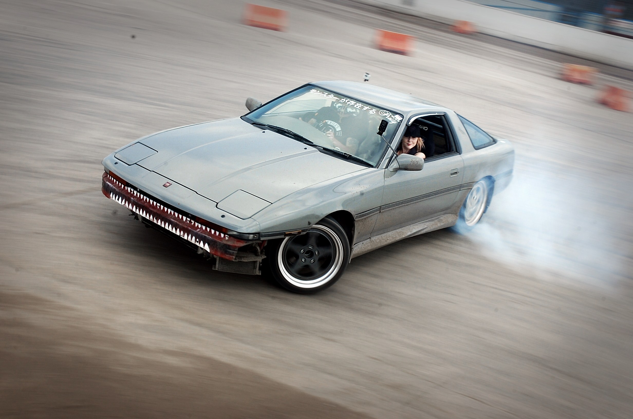 Eine Person in einem Helm fährt ein Auto auf einer Strecke, aus dem Rauch austritt, umgeben von orangefarbenen Gegenständen, mit einem unscharfen Hintergrund, der hohe Geschwindigkeit andeutet.