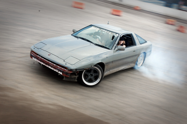 Eine Person in einem Helm fährt ein Auto auf einer Strecke, aus dem Rauch austritt, umgeben von orangefarbenen Gegenständen, mit einem unscharfen Hintergrund, der hohe Geschwindigkeit andeutet.