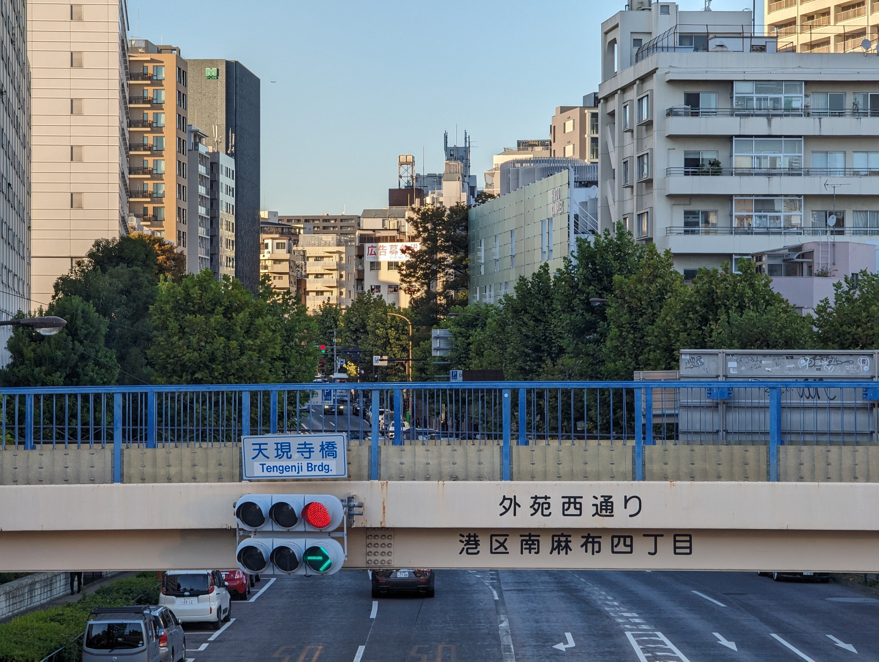 Stadtstraße mit fahrenden Autos, Brücke mit Geländer und Schild, Verkehrsampeln an Pfählen, Laternen, Bäume und Gebäude mit Fenstern unter einem klaren Himmel.