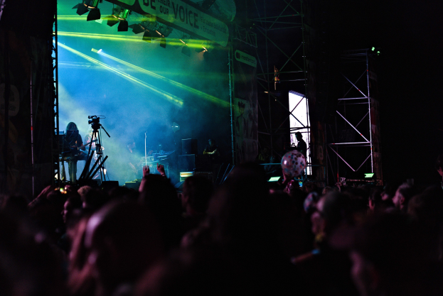 Eine Menge schaut Musikern auf einer hell erleuchteten Bühne bei einem Konzert zu, mit Instrumenten, Mikrofonen und einem Banner im Hintergrund.