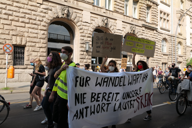 Eine Gruppe von Menschen marschiert auf einer Demonstration gegen die Anti-Kriegsbewegung in Berlin, einige halten Schilder und andere fahren Fahrräder, mit einem Gebäude im Hintergrund, das Fenster, Bögen, Säulen und Skulpturen, Bäume und einen Lichtmast zeigt.