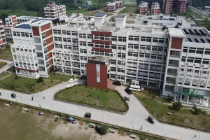 Luftaufnahme eines großen Krankenhausgebäudes mit Solarpaneelen auf dem Dach, umgeben von Bäumen, Gras, Fahrzeugen, Menschen, Strommasten und einer Banner, das ein Solarprojekt anzeigt.