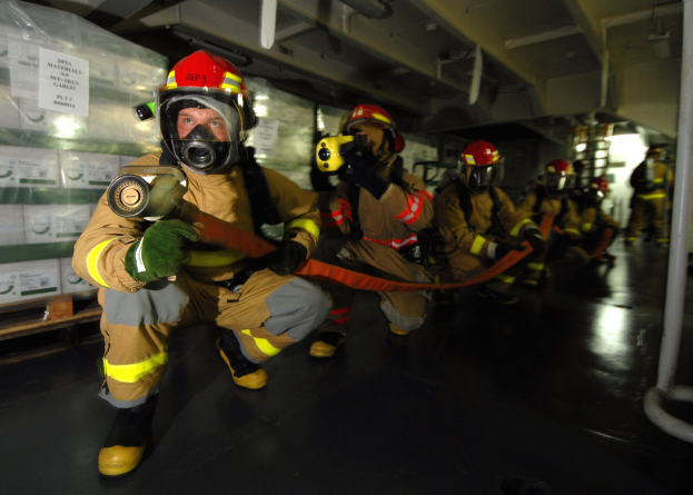 Feuerwehrleute in Schutzausrüstung mit Feuerlöschern, mit beschrifteten Kisten links, einem Pfahl rechts und Überkopfleitungen in einer Schiffsoberdecke.
