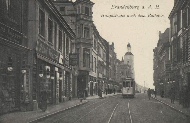 Schwarzes und weißes Foto einer Stadtstraße in Brandenburg, Deutschland, mit einer Straßenbahn, Fußgängern, Gebäuden, Schildern, Straßenlaternen und einem bewölkten Himmel.