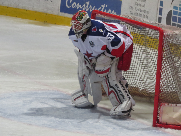 Ein Hockey-Torwart in voller Ausrüstung steht in seinem Tor auf dem Eis, mit einer textbedeckten Wand im Hintergrund.