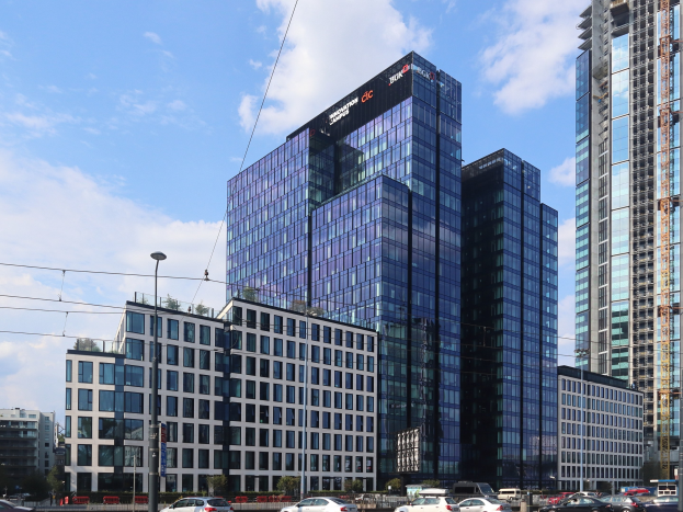 Zentrale der Deutschen Bank in Frankfurt, Deutschland, ein großes mehrstöckiges Gebäude mit zahlreichen Fenstern, umgeben von städtischer Infrastruktur wie Straßenlaternen, Stromleitungen, Fahrzeugen, Grünflächen und einem bewölkten Himmel.