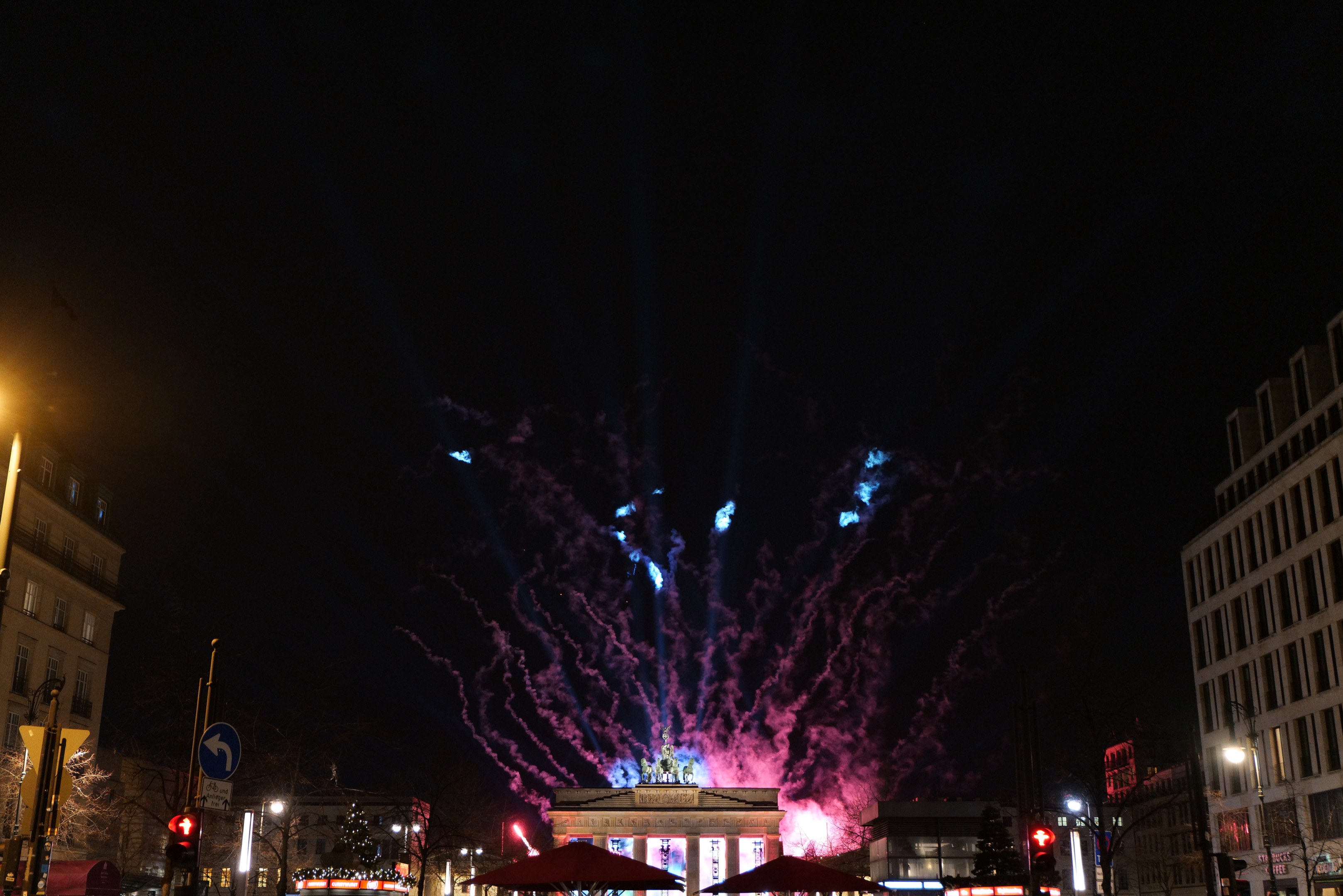 Eine belebte Stadtstraße in Berlin, Deutschland, mit dem Brandenburger Tor im Hintergrund, beleuchtet von Stadtlichtern und Feuerwerken am Nachthimmel.