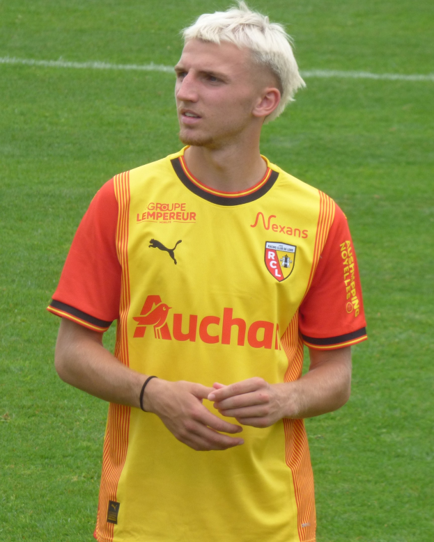 Ein Mann in einem gelb-orangen gestreiften T-Shirt steht selbstbewusst auf einem Fußballfeld und schaut entschlossen nach vorne.