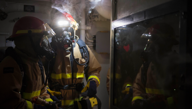Eine Gruppe von Feuerwehrmännern in Gasmasken und Helmen steht vor einer rauchenden Tür, mit Rohren und anderen Gegenständen im Hintergrund, was auf einen begrenzten Raum hinweist.