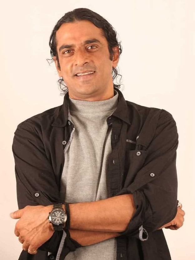 Ein Mann in einem schwarzen und aschfarbenen Kleid mit überschlagenen Armen, der eine Uhr tr├Ągt und gegen einen weißen Hintergrund steht und nachdenklich wirkt.