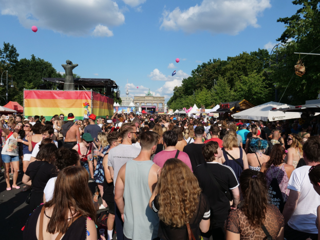 Eine große Menge Menschen geht eine Straße mit Zelten, Bäumen, Pfählen, Lichtern und einer Statue entlang, im Hintergrund Gebäude und ein bewölkter Himmel mit Ballons während des Christopher Street Day in Berlin.