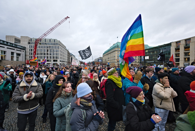 Eine große Gruppe von Menschen vor einem Gebäude mit Fahnen und Schildern, auf denen 'Lgbtq+ rights march in Berlin' steht, einige tragen Mützen und Taschen, im Hintergrund Gebäude, ein Kran und ein bewölkter Himmel.
