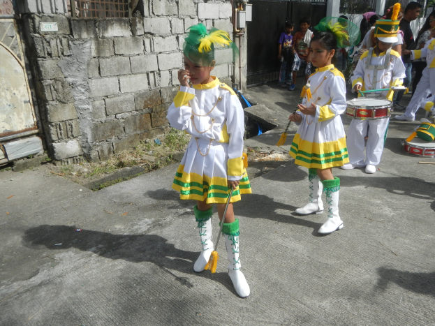 Gruppe junger Mädchen in weißen und gelben Uniformen marschiert, während sie Musikinstrumente spielen und an einer Wand, einem Tor und Bäumen vorbeikommen.