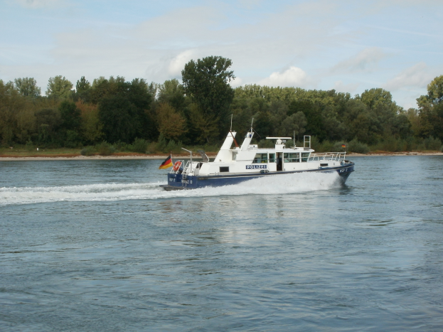 Ein Polizei-Patrouillenboot mit Flaggen fährt einen Fluss hinunter, umgeben von Bäumen unter einem klaren blauen Himmel.