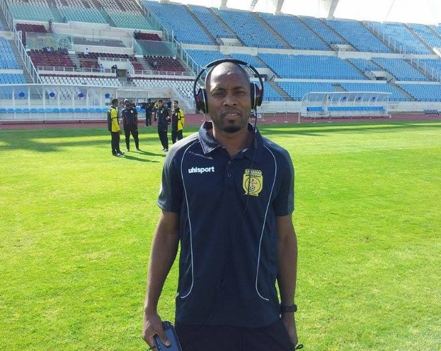 Ein Mann in einem Headset, wahrscheinlich der Trainer der ghanaischen Fußballnationalmannschaft, steht auf einem Fußballfeld in der Nähe eines Stadions und hält ein Objekt, während im Hintergrund Menschen zu sehen sind.