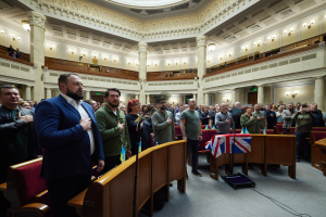 Eine Gruppe von Liberalen, die im House of Commons vor einer Menge, Stühlen, Tischen, Fahnen, einem Mikrofonständer, einem Gerät auf dem Boden, Säulen, Wandlampen und Deckenlampen im Hintergrund stehen.
