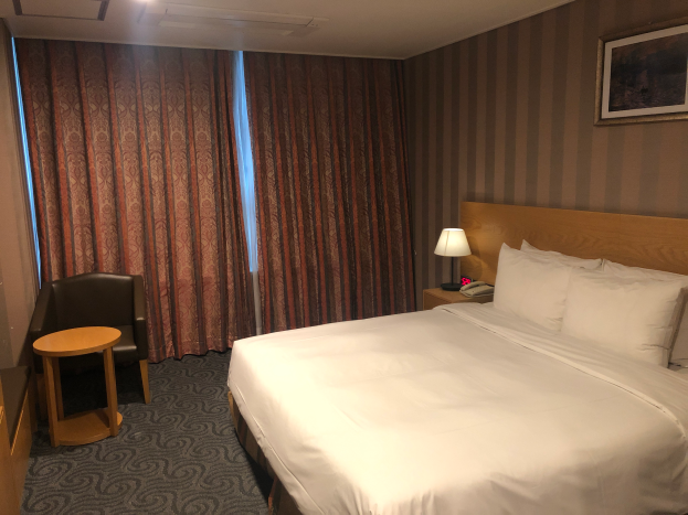 Ein Hotelzimmer mit einem gro├čen Bett mit einer Decke und Kissen, einem Stuhl, einem Tisch mit einem Telefon und einer Lampe, einem Foto auf dem Nachttisch, einem Fenster mit Vorh├Ąngen und einer Decke mit Lampen.