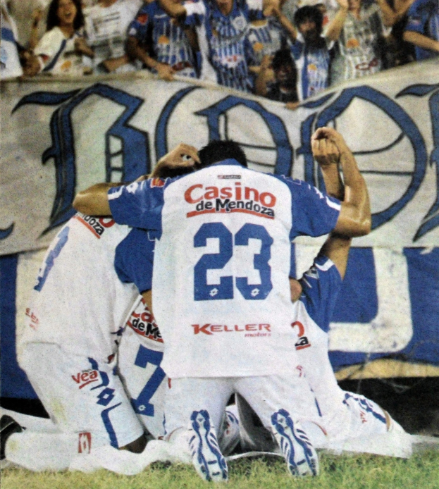 Eine Gruppe von Fußballspielern feiert ein Tor auf dem Feld, mit einem Banner mit Text im Hintergrund und einer jubelnden Menge.