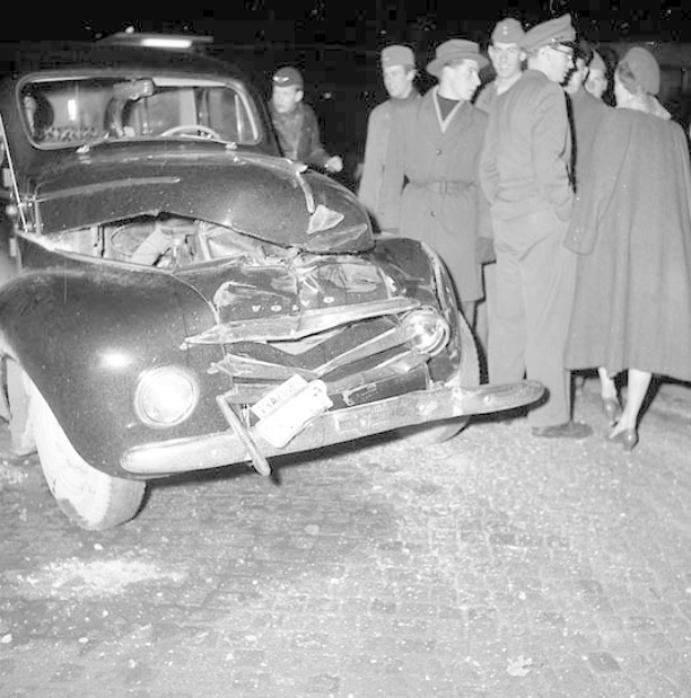 Schwarz-weiß-Bild eines schwer beschädigten Autos am Straßenrand mit eingedellter Motorhaube und zersplitterten Fenstern, umgeben von Menschen, die die Szene begutachten.