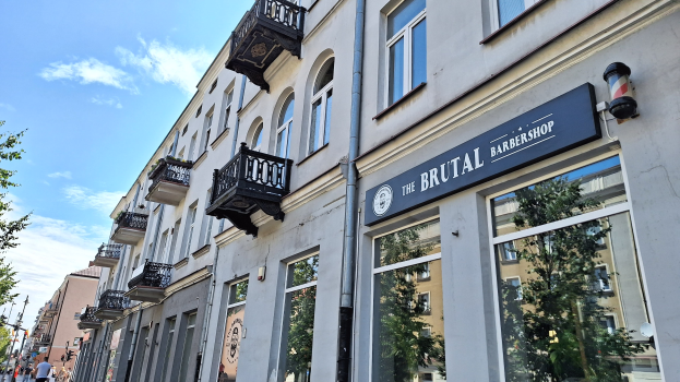 Brutalistischer Friseurladen in Berlin mit Betongebäuden, Fenstern, Geländern, Balkonen und Namensschildern, umgeben von Bäumen, Laternenmasten und Fußgängern an einem bewölkten Tag.