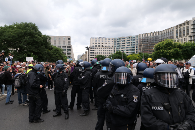 Große Gruppe von Polizeibeamten vor einer Menge während einer Demonstration in Berlin, Deutschland, mit Kameras in der Menge und Bäumen, Gebäuden und einem bewölkten Himmel im Hintergrund.