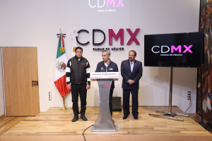 Drei Männer stehen an einem Podium mit einem Mikrofon, flankiert von Fahnen, einer Tafel und einer Wand mit Text, während einer Pressekonferenz in Mexiko.