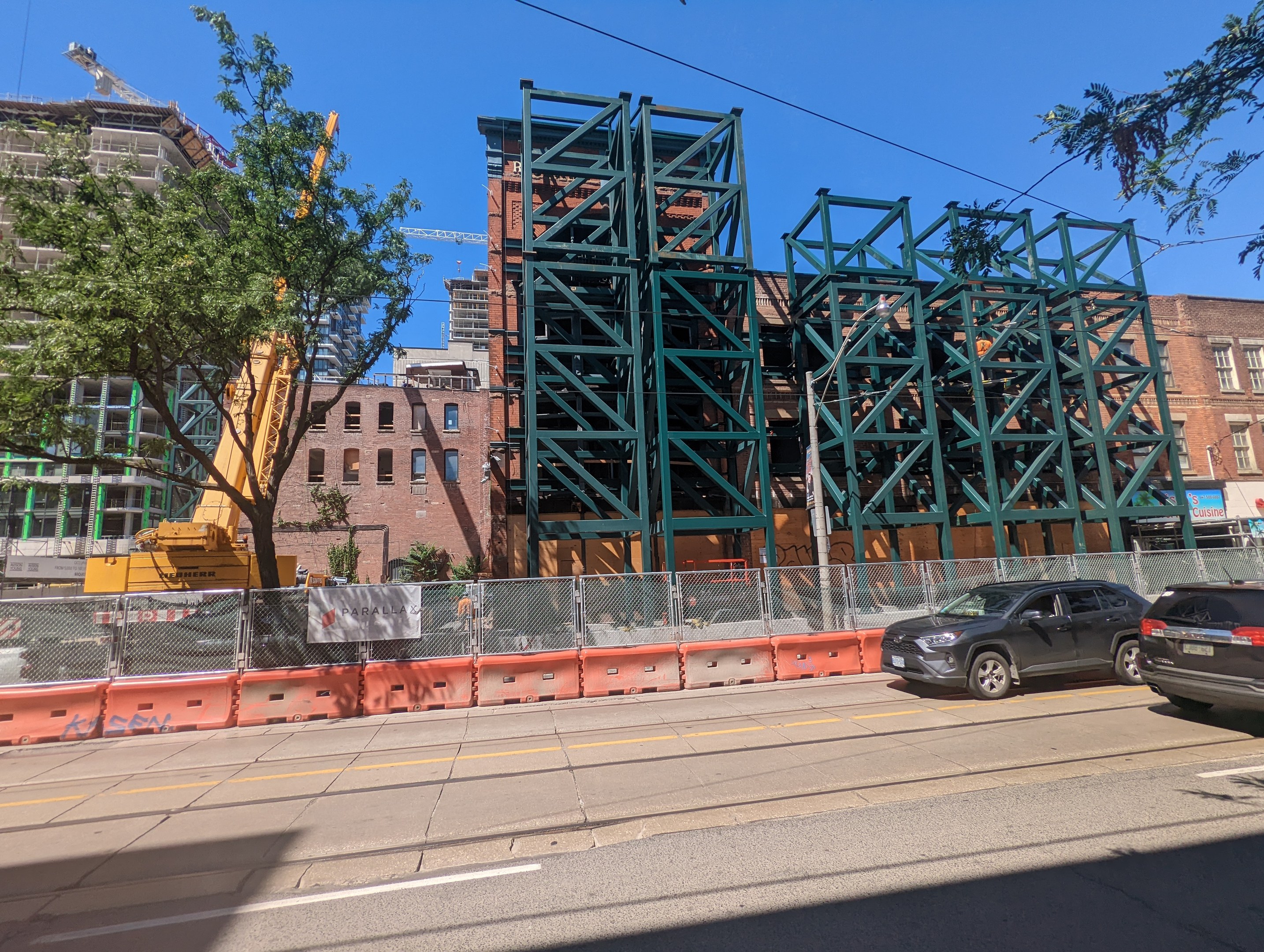 Eine Baustelle mit einer teilweise errichteten Metallbrücke im Vordergrund, parkende Autos an der Straße, ein Zaun, Bäume, Gebäude mit Fenstern, ein Kran und ein Himmel im Hintergrund.