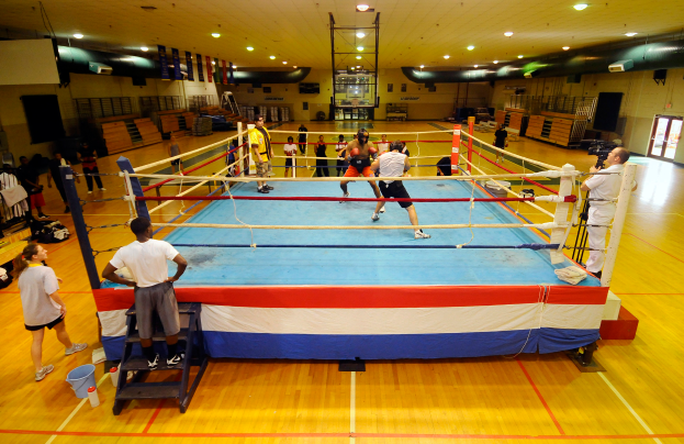 Eine Gruppe von Menschen, die um einen Boxring in einem Turnsaal stehen, mit einigen Personen links und rechts und einer Person mit einer Kamera auf der rechten Seite, unter Deckenlampen.