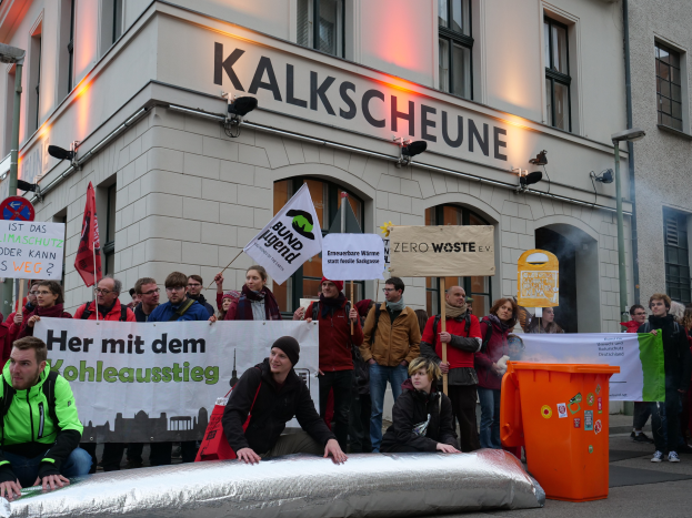 Eine Gruppe von Menschen hält Schilder und Plakate vor einem Gebäude, während zwei Personen im Vordergrund sitzen und ein Mülltonnen auf der rechten Seite zu sehen ist, während einer Protest in Deutschland.
