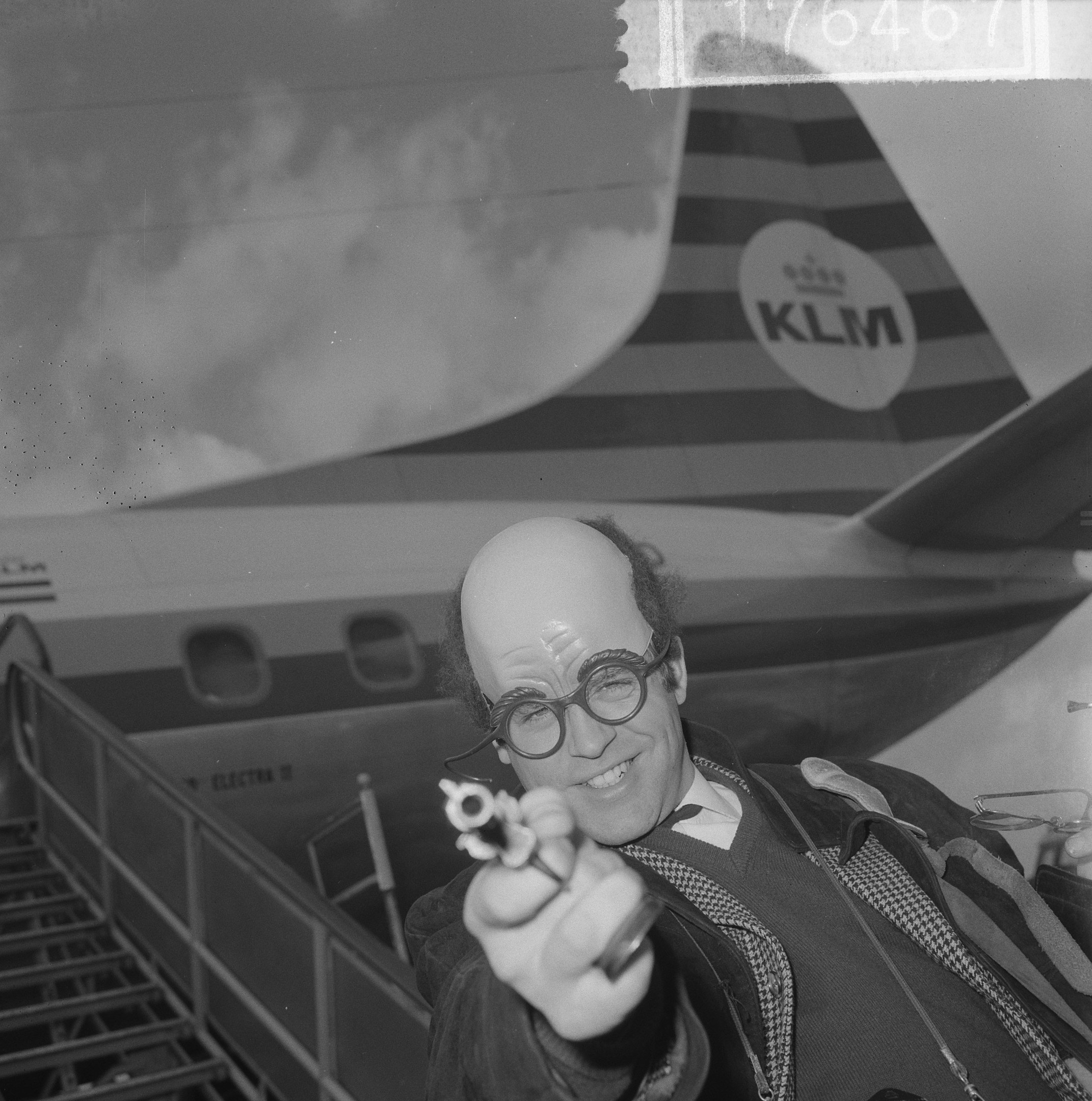 Schwarzes und weißes Bild eines Mannes in einem Anzug und Krawatte, der eine Pistole mit entschlossener Miene hält und vor einem Flugzeug mit ausgebreiteten Flügeln und brüllenden Motoren steht.