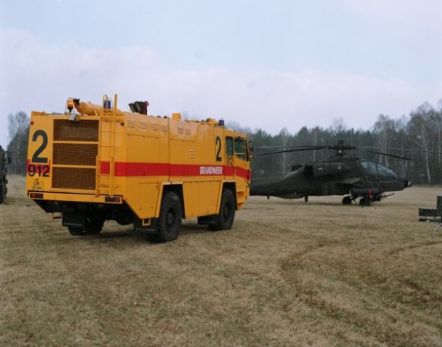 Ein gelber Feuerwehrauto neben einem schwarzen Hubschrauber auf einer grünen Wiese geparkt, mit Bäumen im Hintergrund und einem klaren blauen Himmel darüber.
