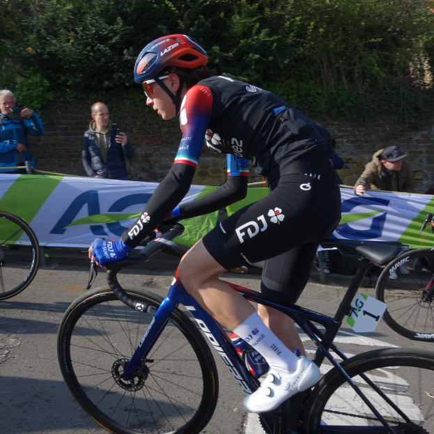 Die professionelle Radsportlerin Katie Archibald fährt mit Helm, Brille, Handschuhen und Schuhen auf einem Fahrrad die Straße entlang, umgeben von einer Menge und einem Banner.