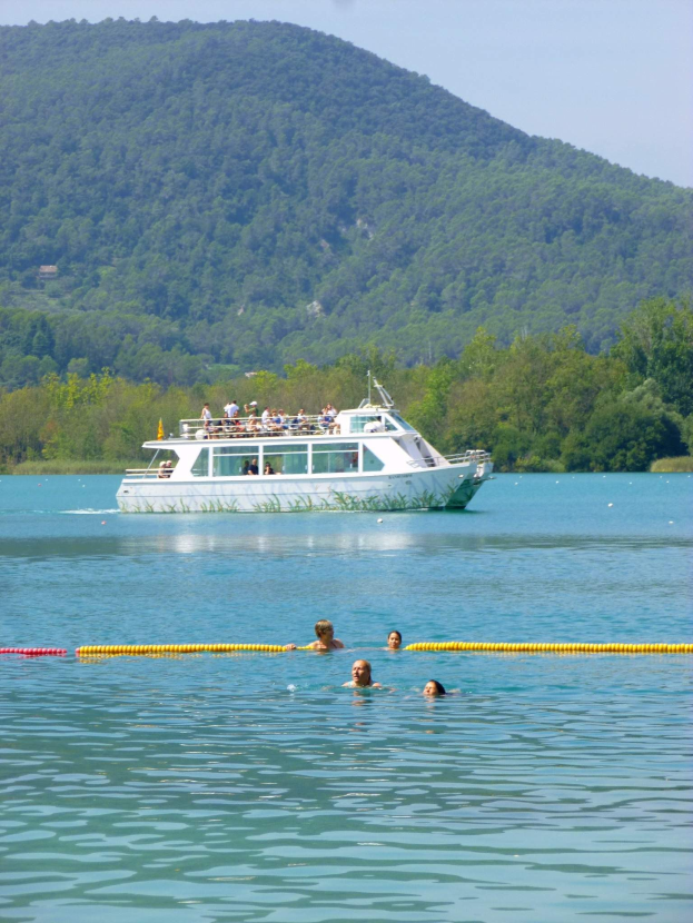 Gruppe von Menschen, die in der Nähe eines Bootes schwimmen, mit einem gelben Rohr im Vordergrund, Bäumen und einem Berg im Hintergrund unter einem klaren blauen Himmel.