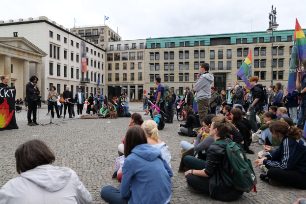 Menschen sitzen auf dem Boden vor einer Menge mit Fahnen und Spruchbändern bei einer Anti-Schwulen-Demonstration in Berlin, mit einem Redner, einer Statue und Gebäuden im Hintergrund.