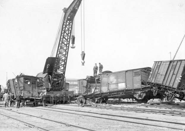 Schwarzes und weißes Bild von einer Zugkatastrophe mit einem Kran, der einen Zugwagen auf die Schienen hebt, mehrere Menschen in der Nähe und im Hintergrund Gebäude und einen klaren Himmel.