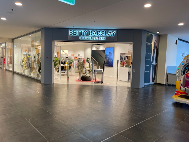 Das Innere eines Betty Barclay-Ladens in einem Einkaufszentrum mit Schaufensterpuppen in Kleidern, hängenden Kleidern, Tischen, Pflanzen und anderen Gegenständen sowie einem Spielzeugfahrzeug auf der rechten Seite und einer Leuchtschrift mit der Aufschrift "Betty Barclay Selected Brands" oben.