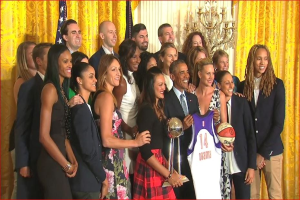 Präsident Obama und First Lady Michelle Obama posieren mit dem Damen-Basketball-Team im Oval Office des Weißen Hauses, halten einen Basketball, Pokal und lächeln in der Nähe einer Flagge, Vorhänge und eines Kerzenleuchters.