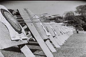 Schwarz-weisses Foto eines Bogenschiess-Ziels mit Pfeilen in der Mitte, umgeben von Bäumen und einer Person im Hintergrund, mit dem Boden unten sichtbar.