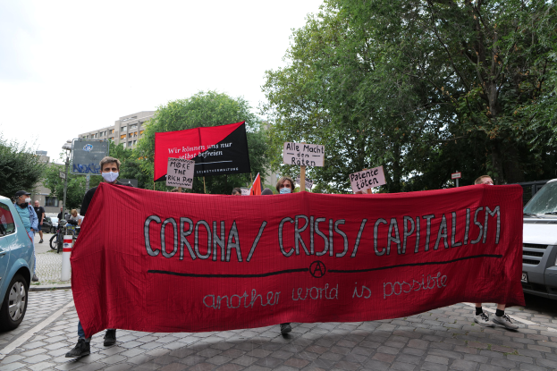 Eine Gruppe von Menschen marschiert eine Straße entlang und hält ein rotes Banner mit der Aufschrift 'Corona-Krise/Kapitalismus', mit parkenden Fahrzeugen, Gebäuden und Bäumen im Hintergrund unter einem klaren blauen Himmel.