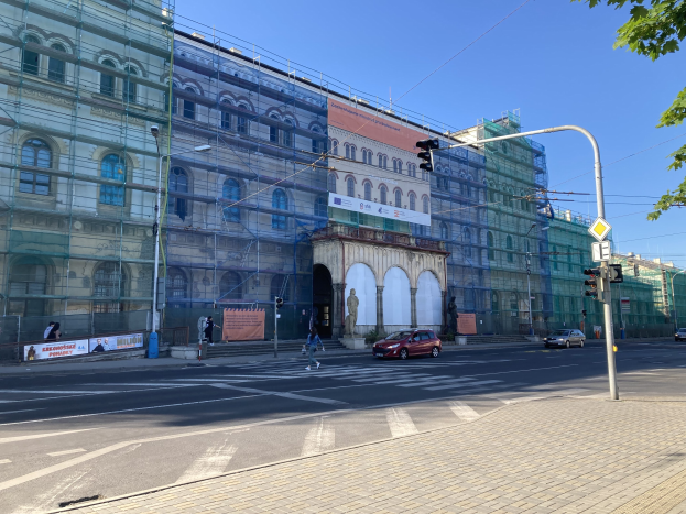 Großes Gebäude mit Gerüst, das renoviert wird, umgeben von Fahrzeugen, Passanten, Ampeln, Straßenschildern, Pfählen, Bäumen und einem klaren blauen Himmel.