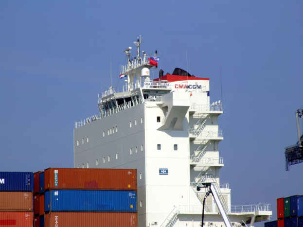 Ein großes Containerschiff liegt im Wasser mit zahlreichen Containern im Vordergrund und einem Kran auf der rechten Seite, unter einem klaren Himmel.