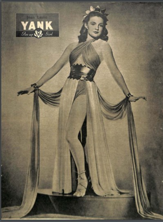 Ein Schwarz-Weiß-Foto der Schauspielerin Joan Leslie in einem langärmligen, hochgeschlossenen Kleid mit einer 1940er-Jahre-Frisur und Make-up, die selbstbewusst auf einer Bühne steht.